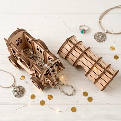 Assembled laser-cut wooden wagon puzzle box with silver chains inside, next to its roof piece, on a white wooden surface with fairy lights