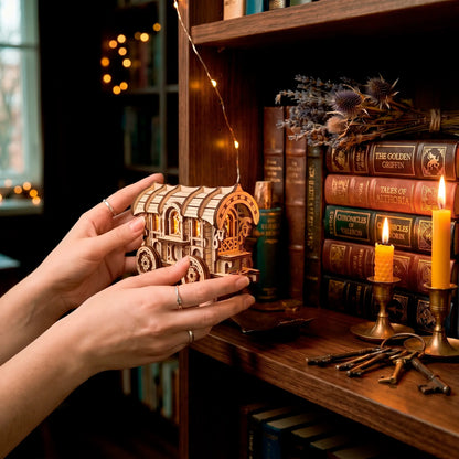 Hands hold the lit laser-cut wooden wagon model in a dim, cozy library filled with vintage books, candles, and old keys.