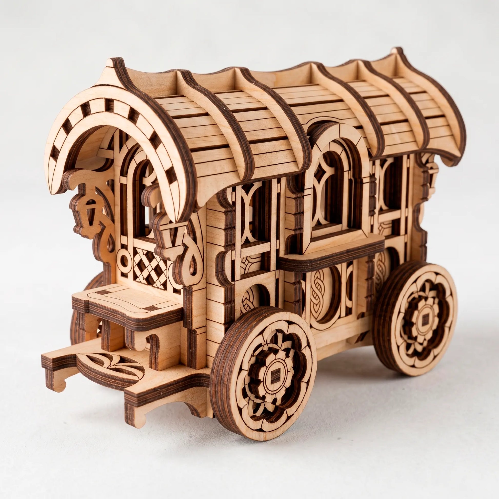 A laser-cut wooden vintage caravan model with intricate window lattice and spoked wheels against a neutral background.