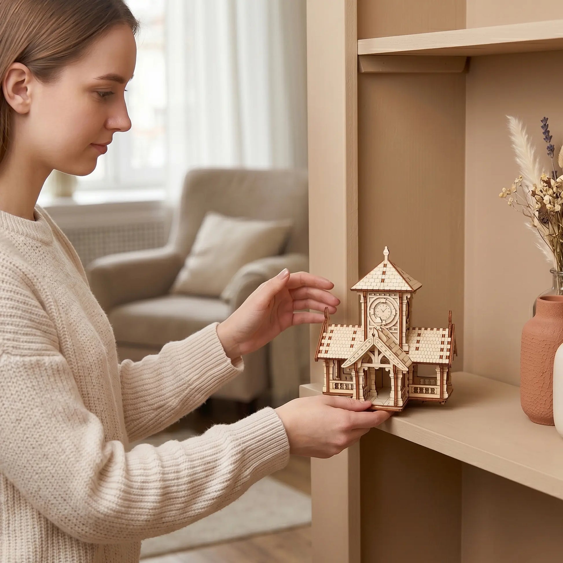 A woman placing a miniature laser cut plywood clock tower house model onto a beige bookshelf in a cozy room setting.