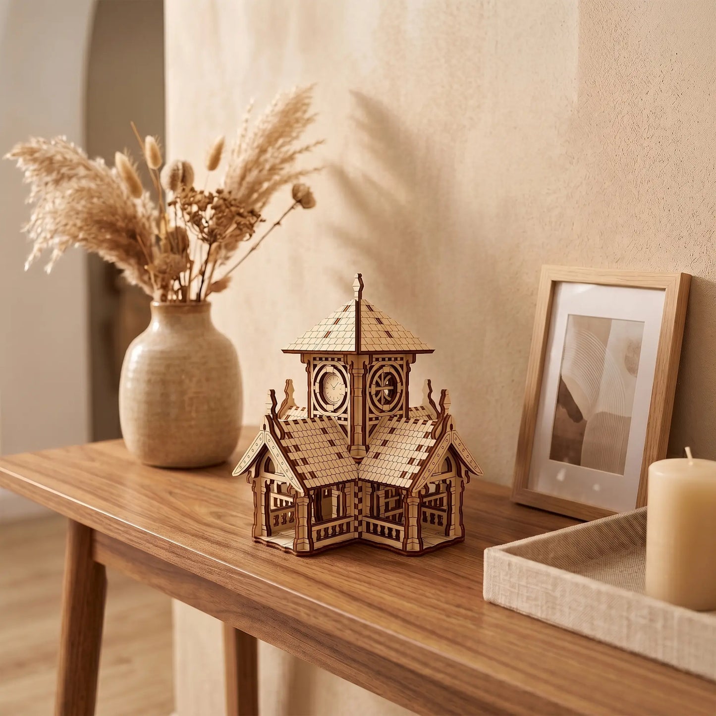 Miniature clock tower house of laser cut plywood staged on a wooden console table with a pampas grass vase.