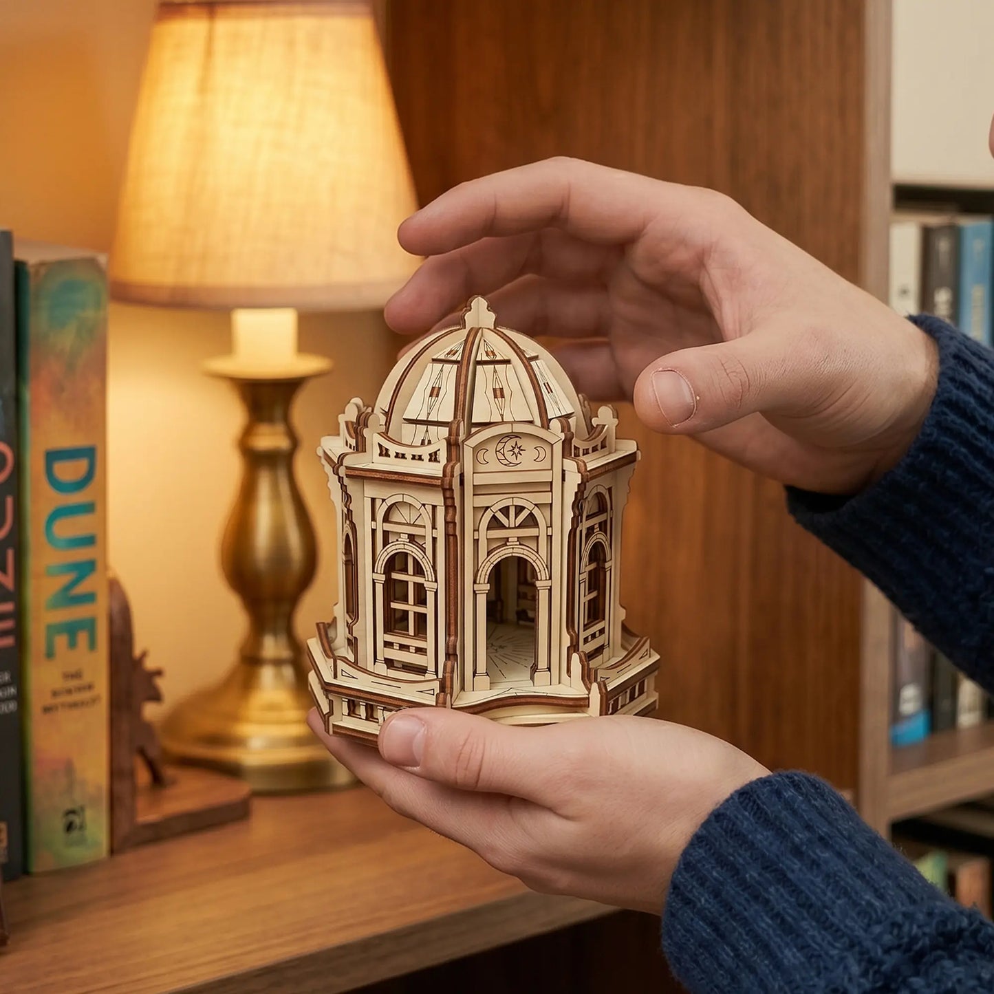Hands holding a small laser cut plywood moon temple miniature on a desk near a cozy lamp and Dune books.