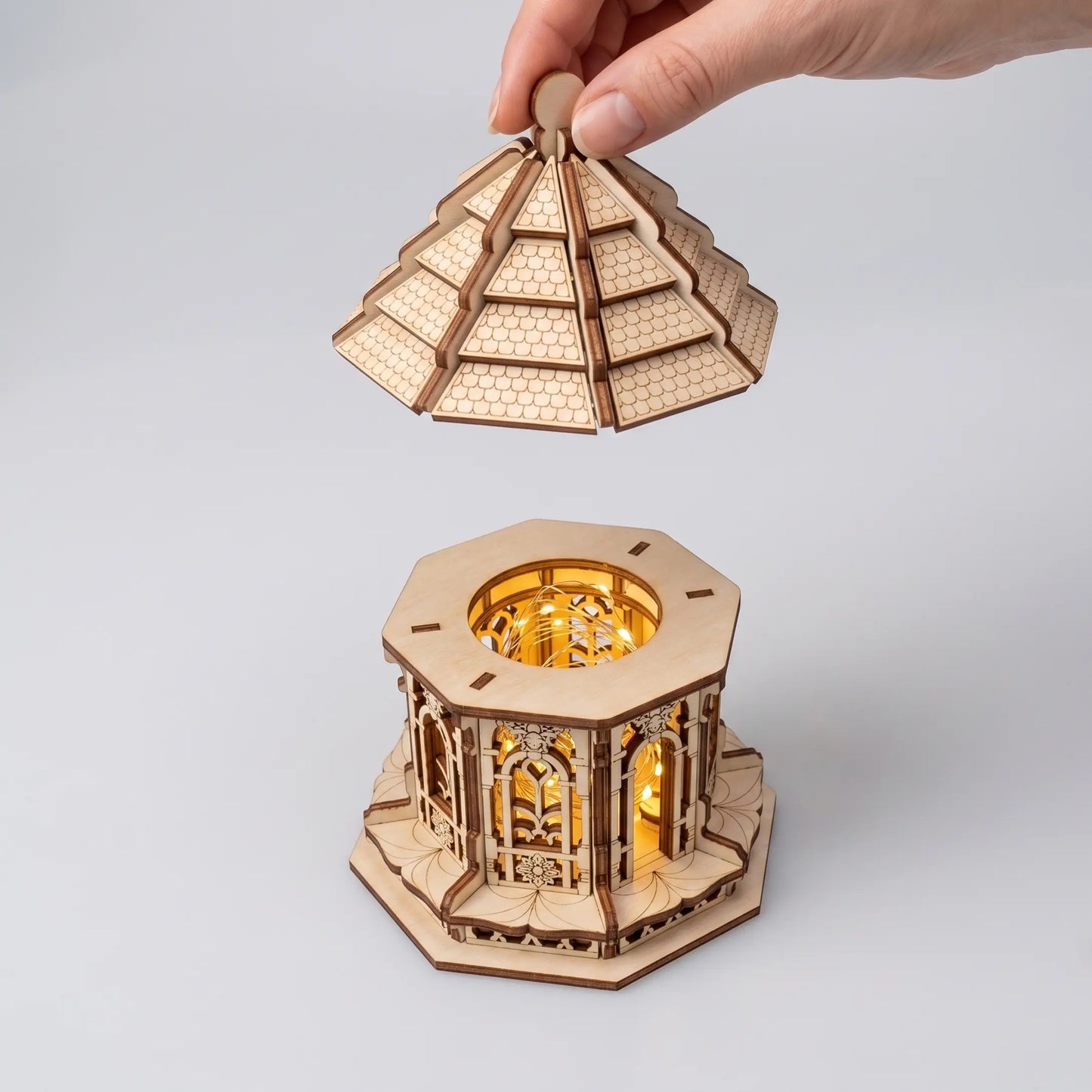 A wooden laser cut fairy house model showing a hand lifting the top pyramid lid of a hexagonal gazebo structure, with the illuminated interior visible below on a light gray background.