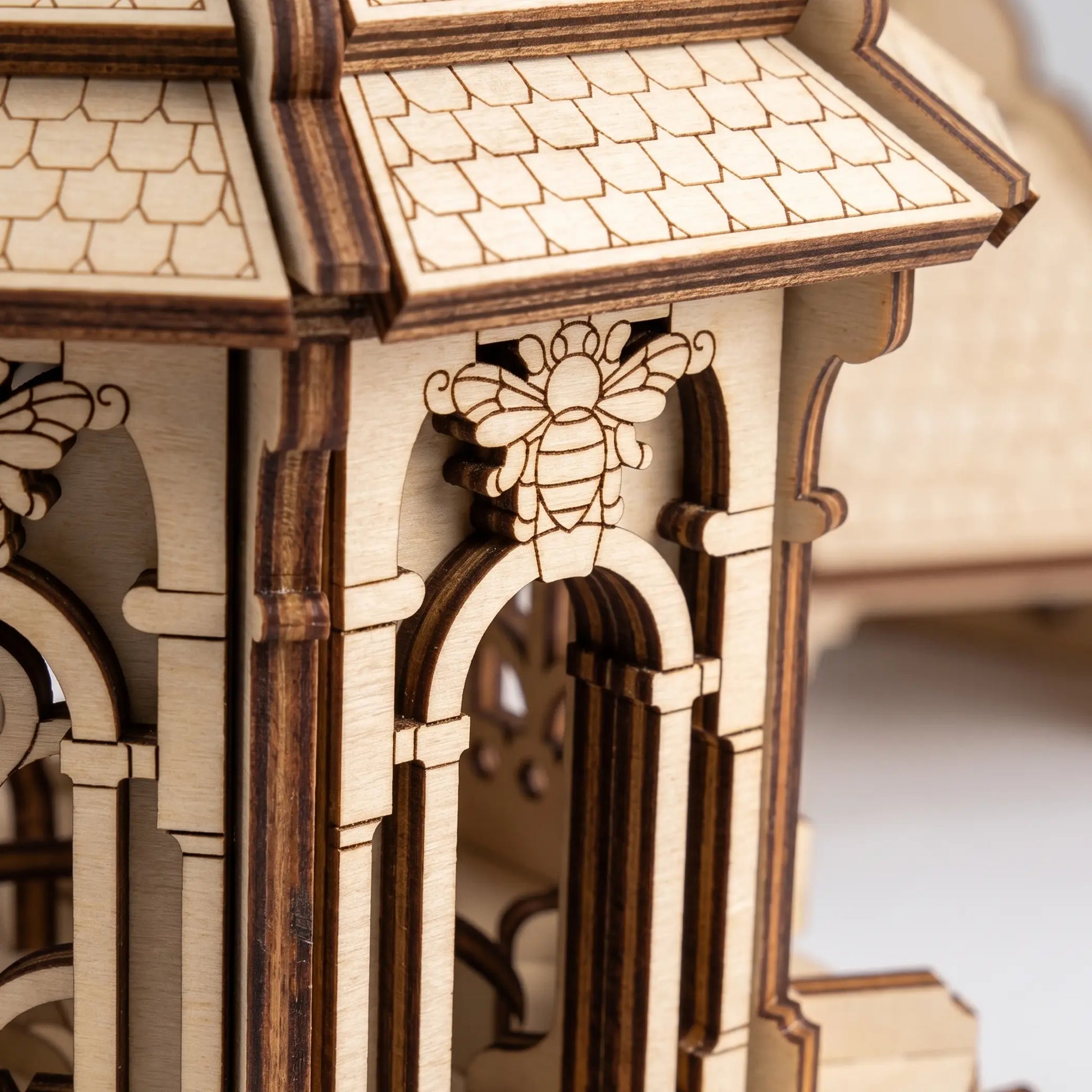 Close-up of a laser-cut wooden model structure with arched windows, a tiled roof, and a carved bee detail on the front.