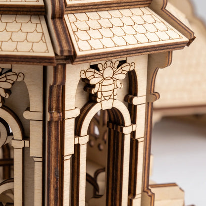 Close-up of a laser-cut wooden model structure with arched windows, a tiled roof, and a carved bee detail on the front.