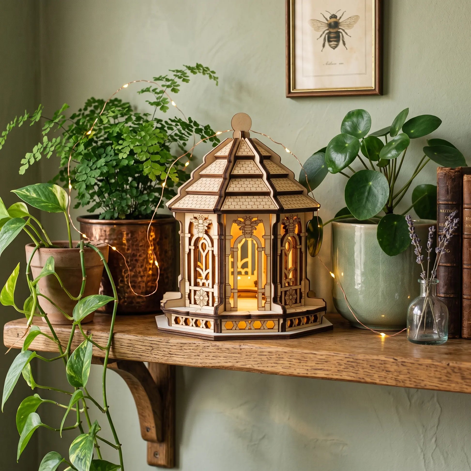 A wooden laser cut fairy house shaped lantern with a glowing warm light inside, displayed on a rustic shelf alongside plants, books, and string lights in a cozy indoor setting.