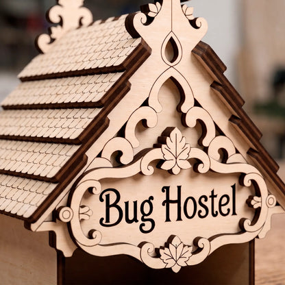 Close-up of the "Bug Hostel" laser-cut plywood sign on the roof gable, featuring ornate leaf carvings and dark burnt edges.
