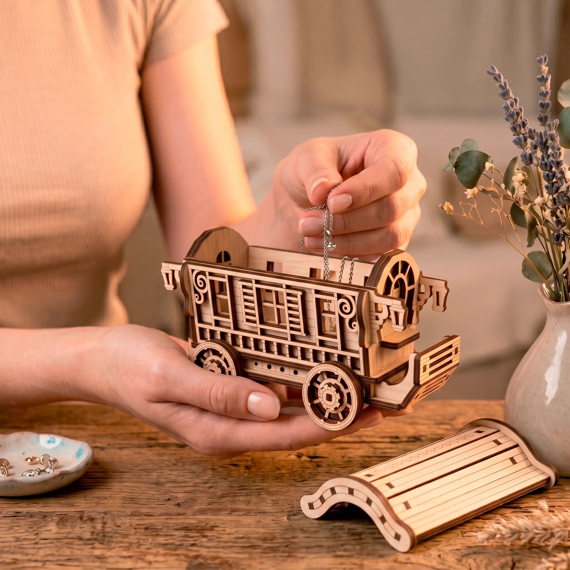 A person holding an open laser-cut plywood fairy wagon organizer while carefully placing a delicate silver necklace inside it.
