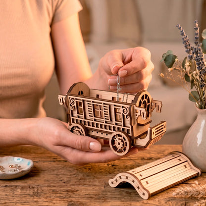 A person holding an open laser-cut plywood fairy wagon organizer while carefully placing a delicate silver necklace inside it.