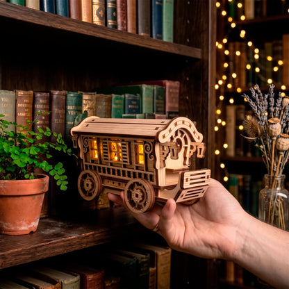 A person holding a glowing laser-cut plywood fairy nomad wagon illuminated from within against a dark bookshelf backdrop.