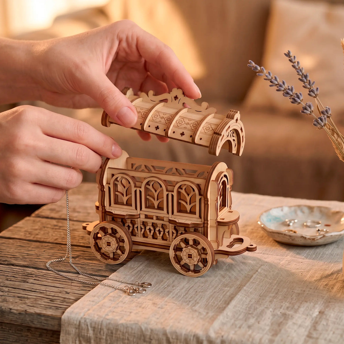 A person carefully lifting the decorative roof lid off an open laser-cut plywood fairy cart jewelry box on a wooden table