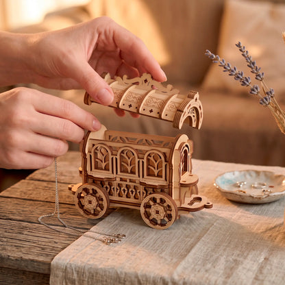 A person carefully lifting the decorative roof lid off an open laser-cut plywood fairy cart jewelry box on a wooden table