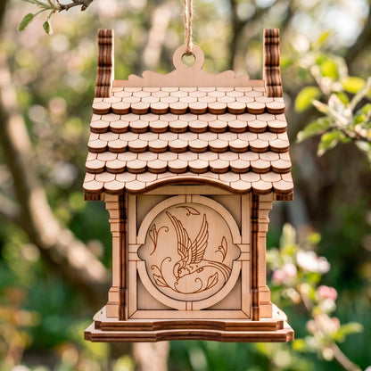 A natural laser-cut plywood birdhouse with shingled roof hanging by a sturdy twine string outdoors against a blurred garden.