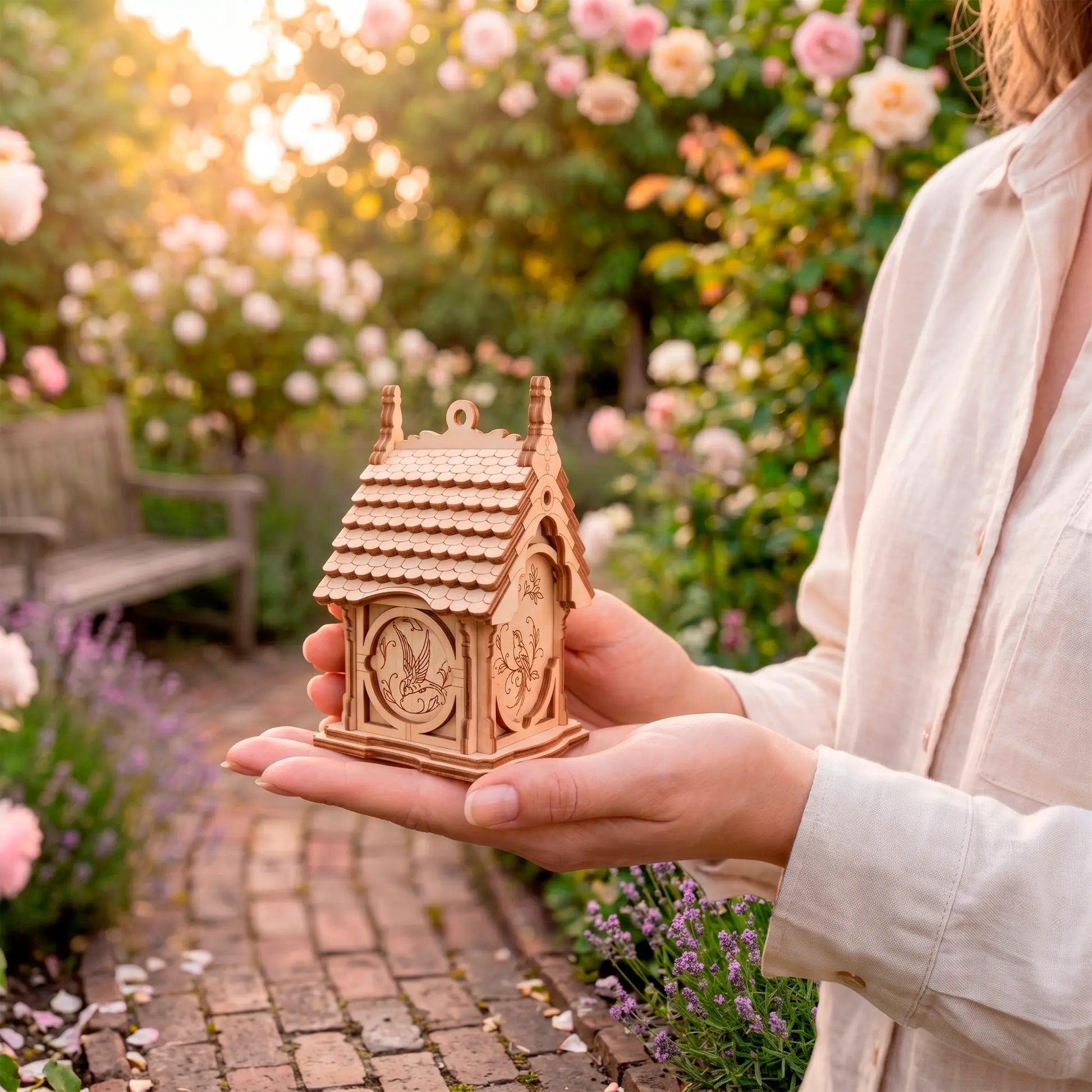 A person carefully holding a small natural laser-cut plywood birdhouse in their open hands in a blooming garden at sunset.
