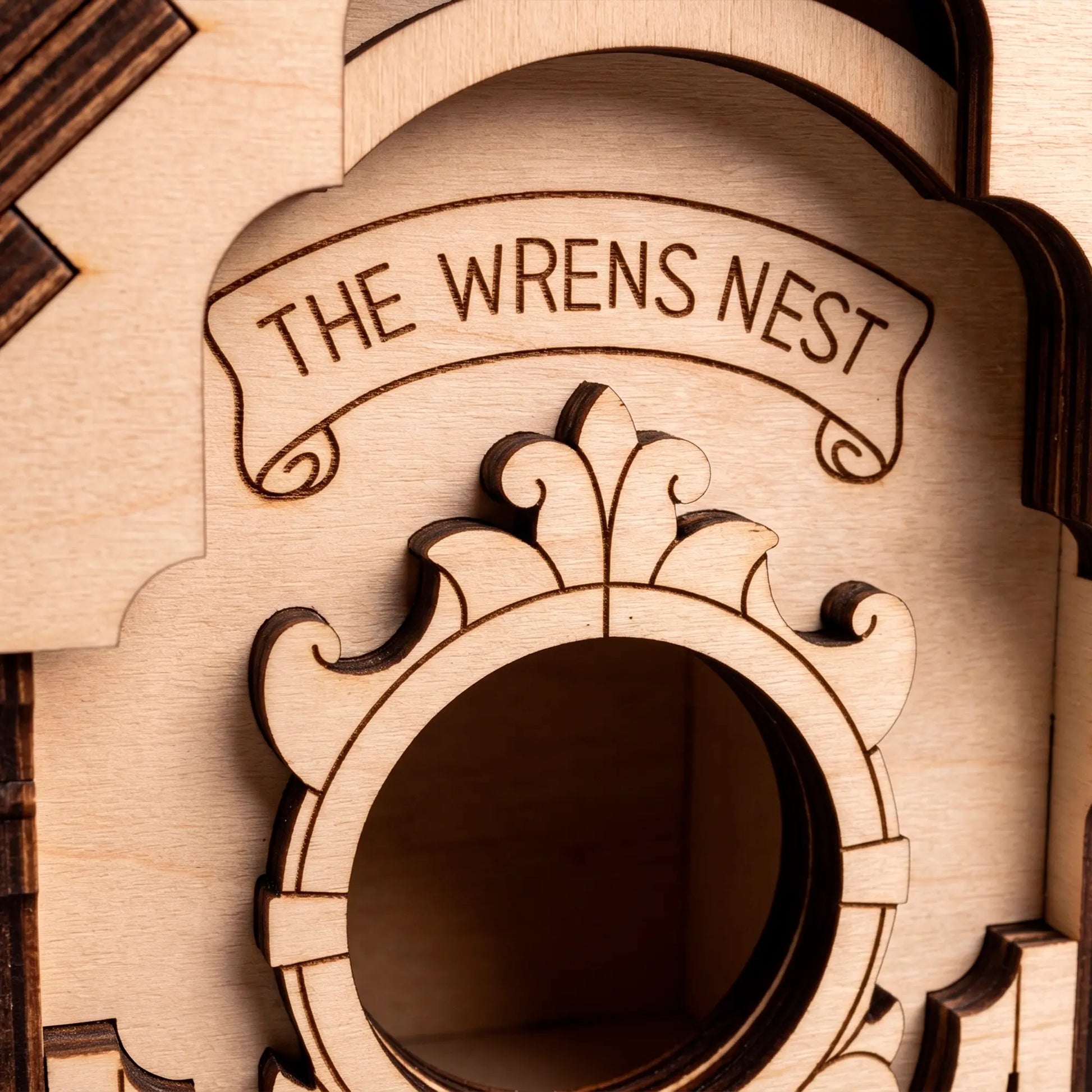 A crisp macro close-up of a laser-cut plywood birdhouse facade featuring the engraved text 'The Wrens Nest' above the entry