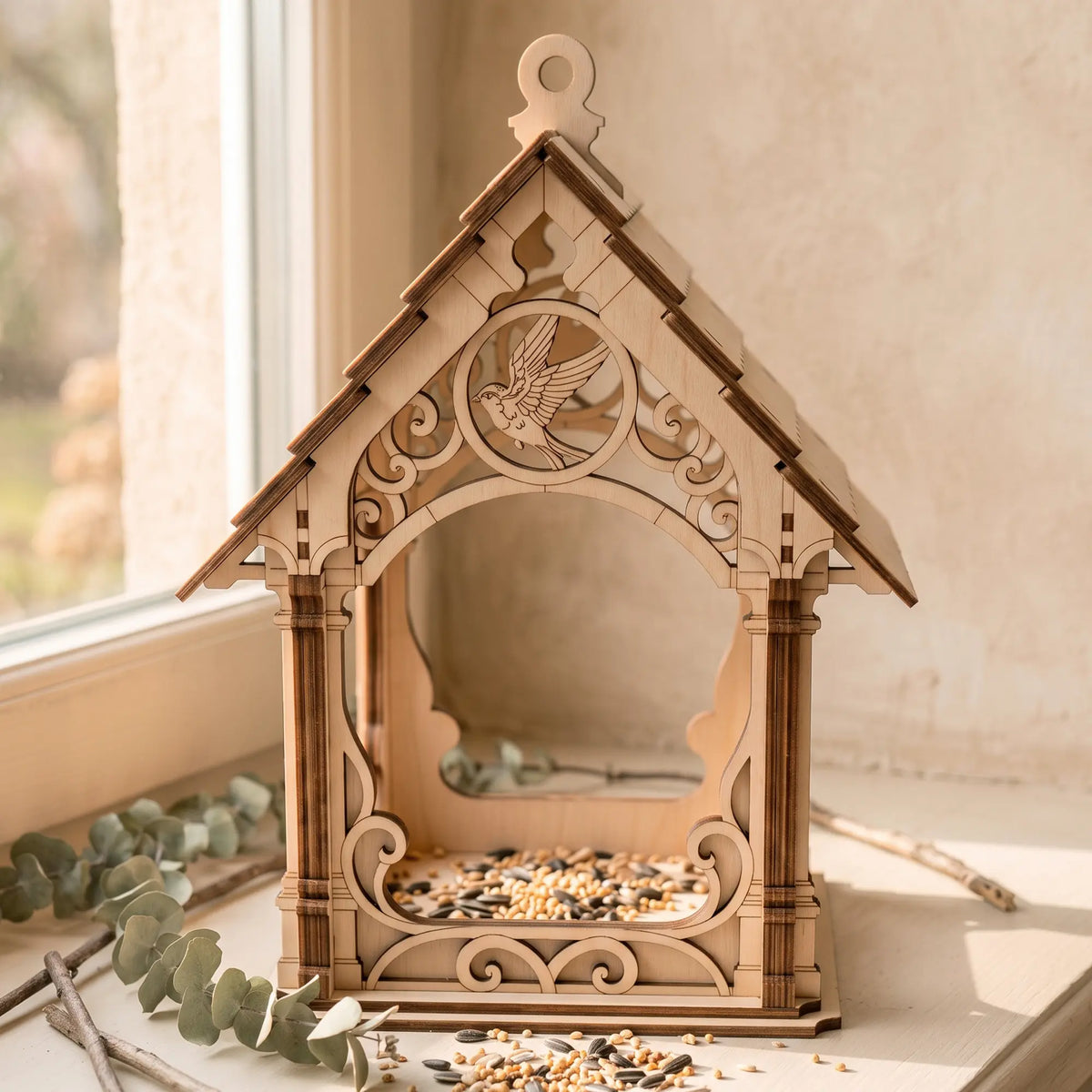 Laser cut plywood bird feeder house filled with seeds resting on sunlit indoor windowsill next to eucalyptus leaves.