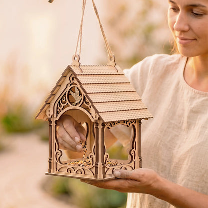 Woman holds hanging laser cut plywood bird feeder house with twine rope, scattering seed mix inside outdoors