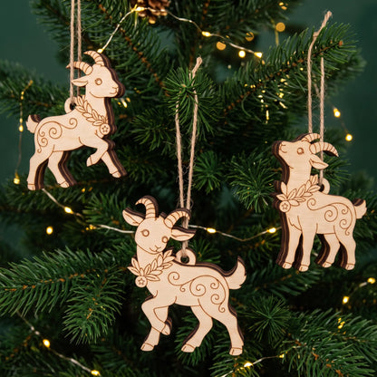 Three laser cut plywood goat ornaments hanging by twine strings on green Christmas tree branches with warm fairy lights.