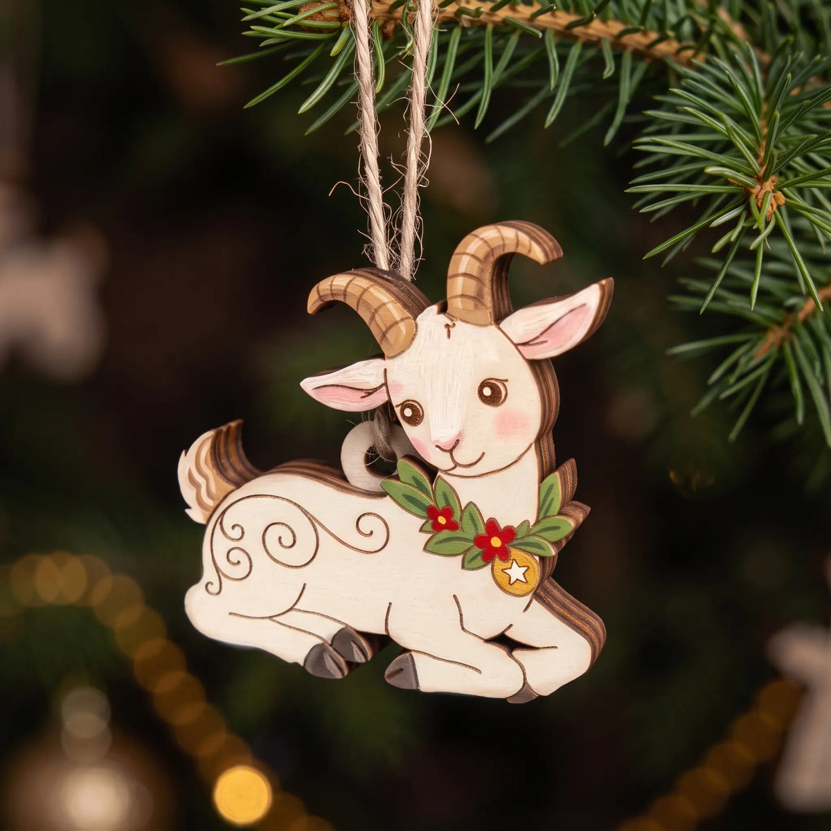 Close-up of painted laser cut plywood resting goat ornament with horns and floral wreath hanging on Christmas tree branch.