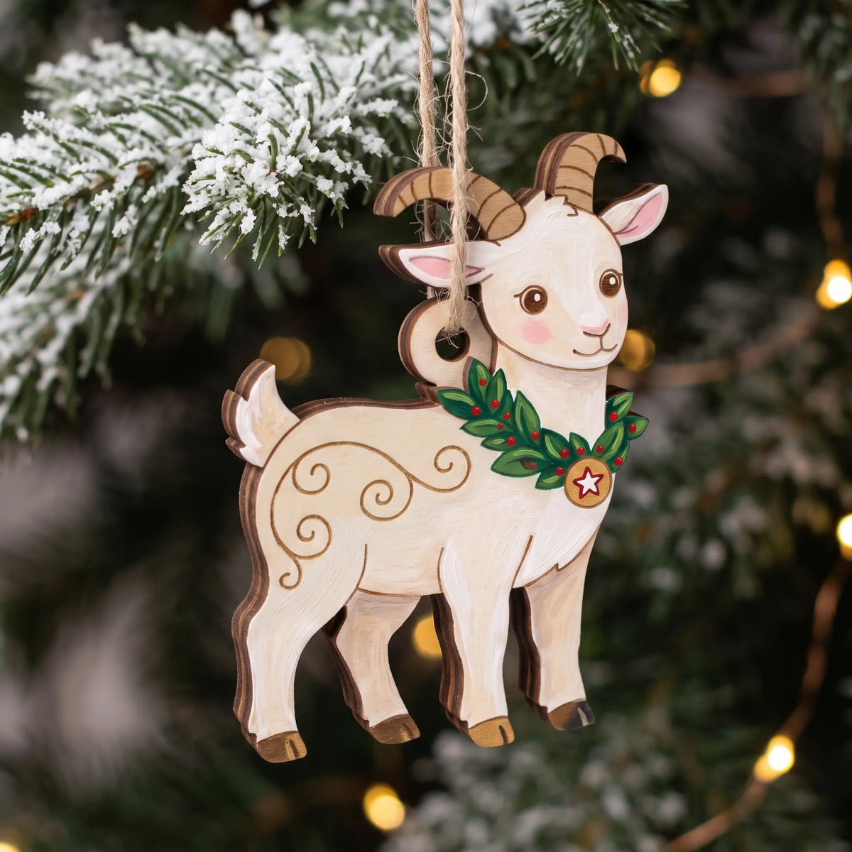Painted laser cut plywood goat ornament wearing wreath collar hanging on snow-dusted Christmas pine branch with bokeh.