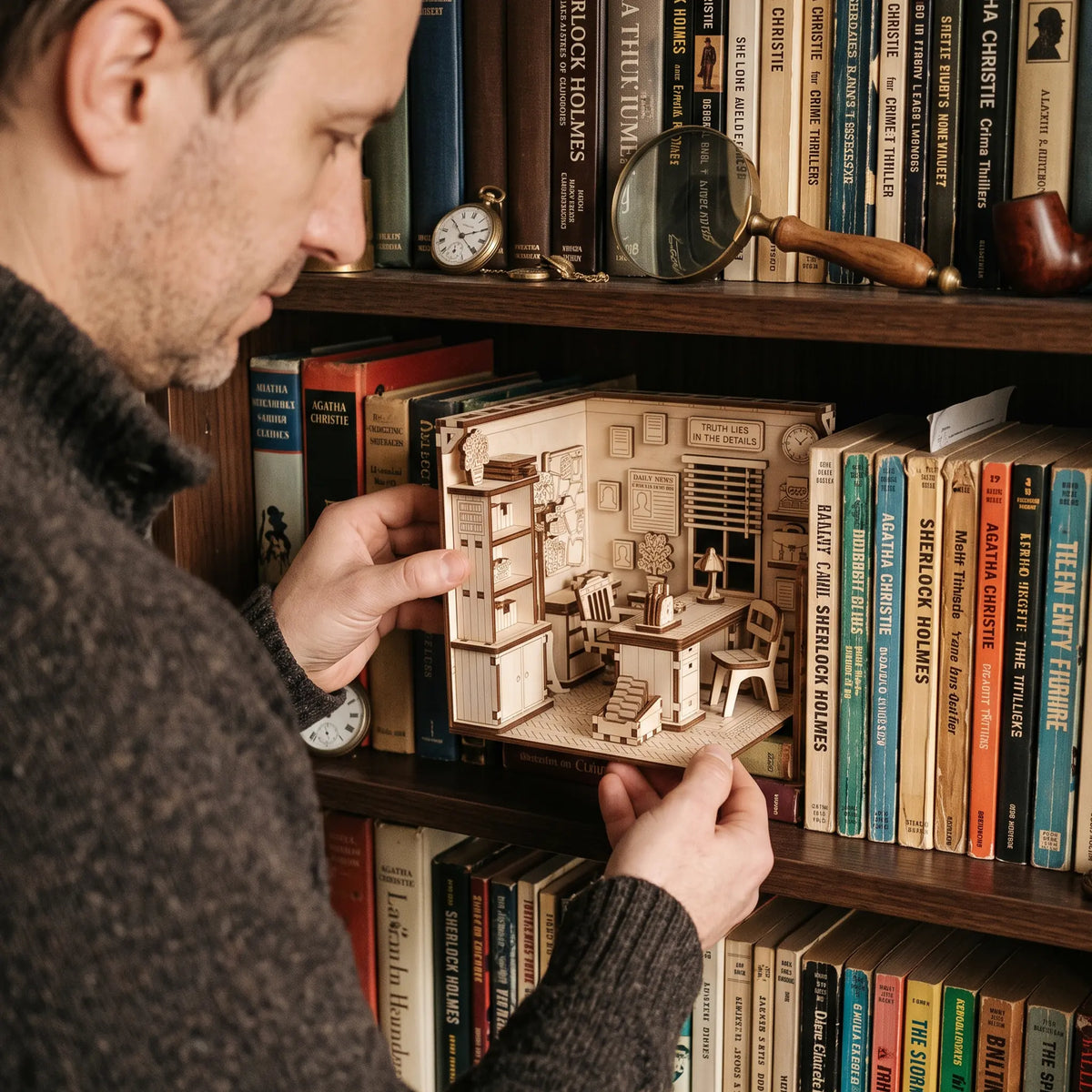 Man placing a laser cut plywood detective office book nook onto a dark bookshelf between vintage Sherlock Holmes novels.