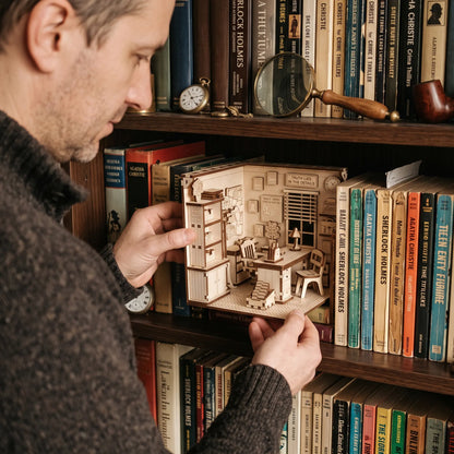 Man placing a laser cut plywood detective office book nook onto a dark bookshelf between vintage Sherlock Holmes novels.