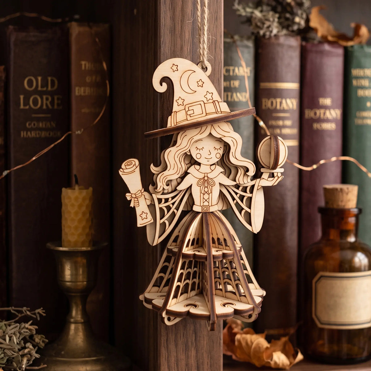 Laser cut plywood witch ornament hanging near old lore books, candle, and vintage apothecary bottle.