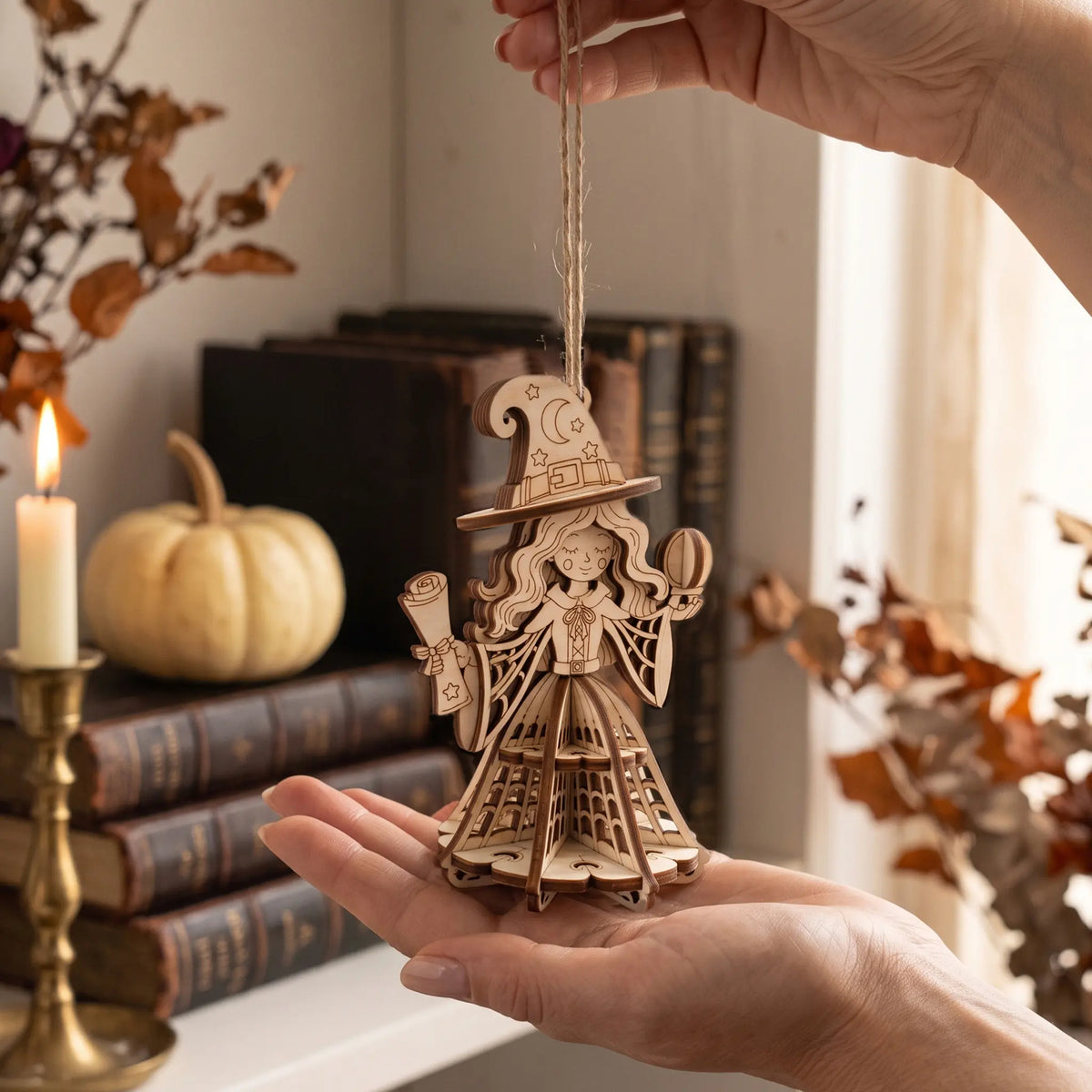 Hand holding a 3D laser cut plywood Halloween witch pendant over candle and white pumpkin on desk.