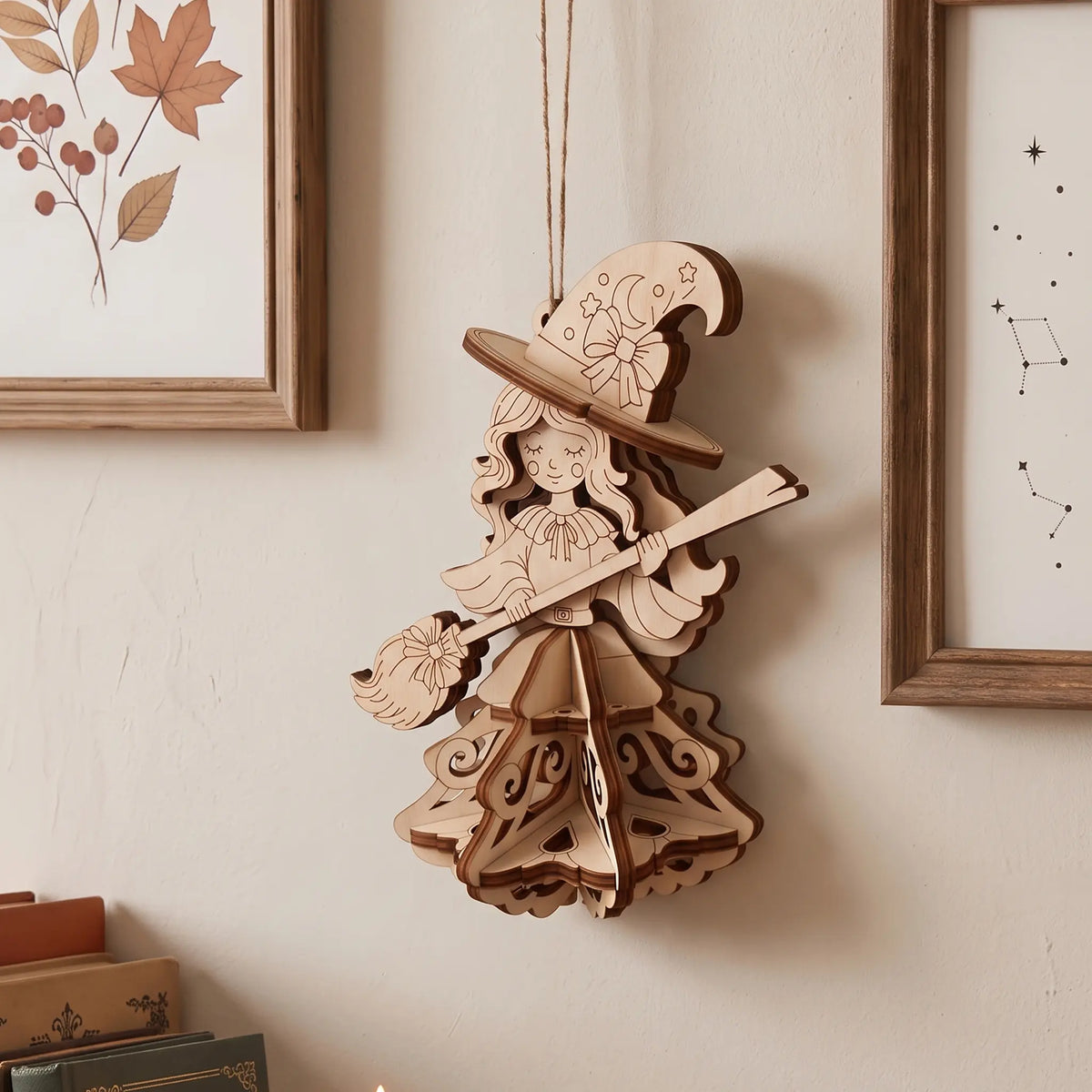 Laser cut plywood witch ornament hanging on a light wall between two wooden photo frames above vintage books on a shelf.