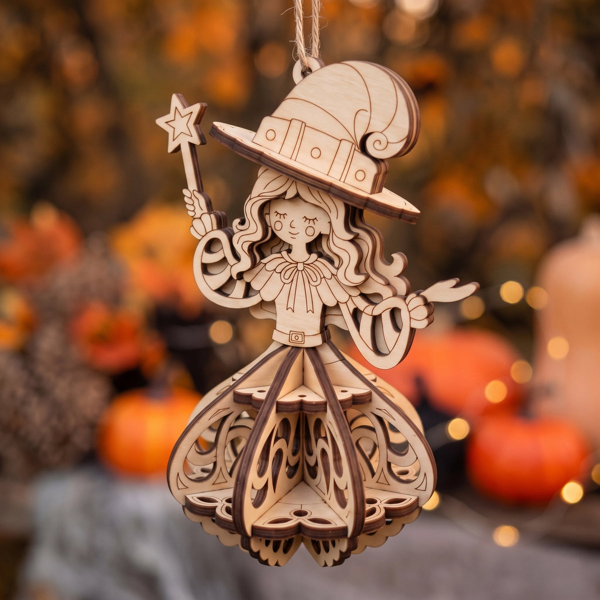 Hanging laser cut plywood witch ornament holding a magic star wand against a warm blurred autumn foliage background.