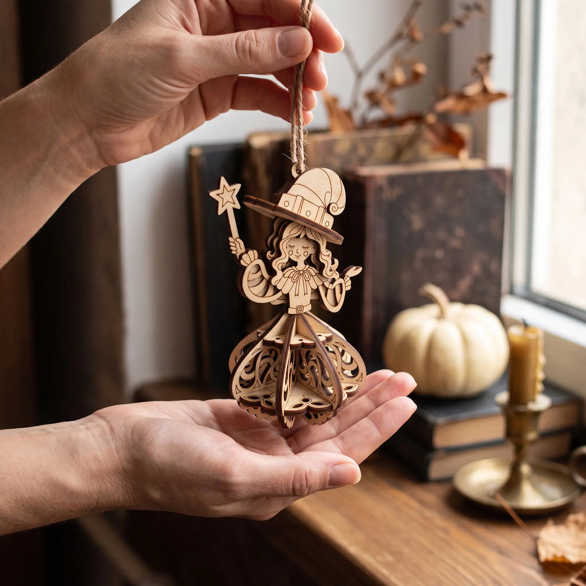 Woman holding laser cut plywood witch ornament in palm, showing scale near old vintage books and autumn windowsill.