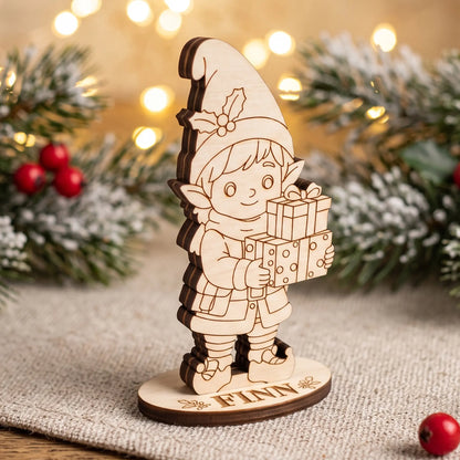 Unpainted laser cut plywood elf Finn figurine holding gift boxes on a wooden stand by pine branches.