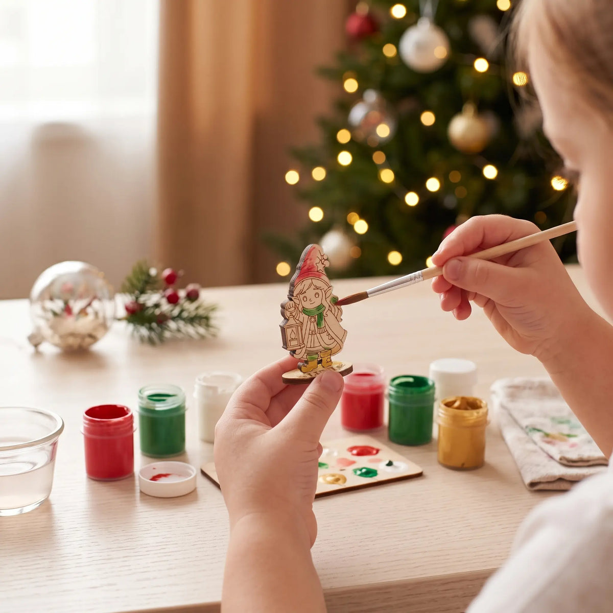 Child hands painting a laser cut plywood elf figurine with acrylic paint and brush at Christmas craft desk.