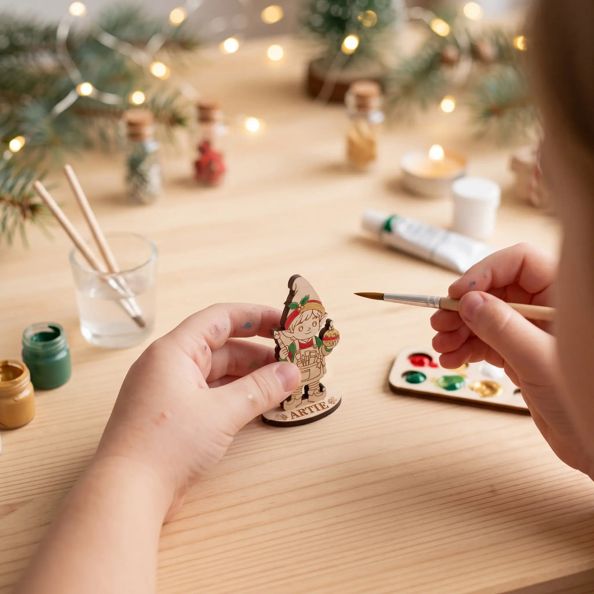 Hand painting laser cut plywood artist elf Artie figurine with brush and paint pots on light wooden desk.