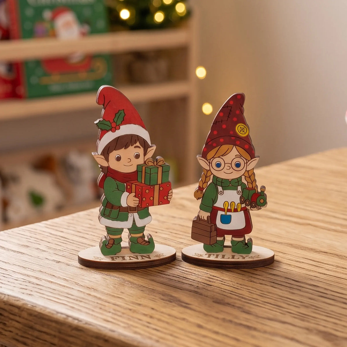 Painted laser cut plywood standing elves Finn and Tilly on wooden desk in front of cozy Christmas lights.