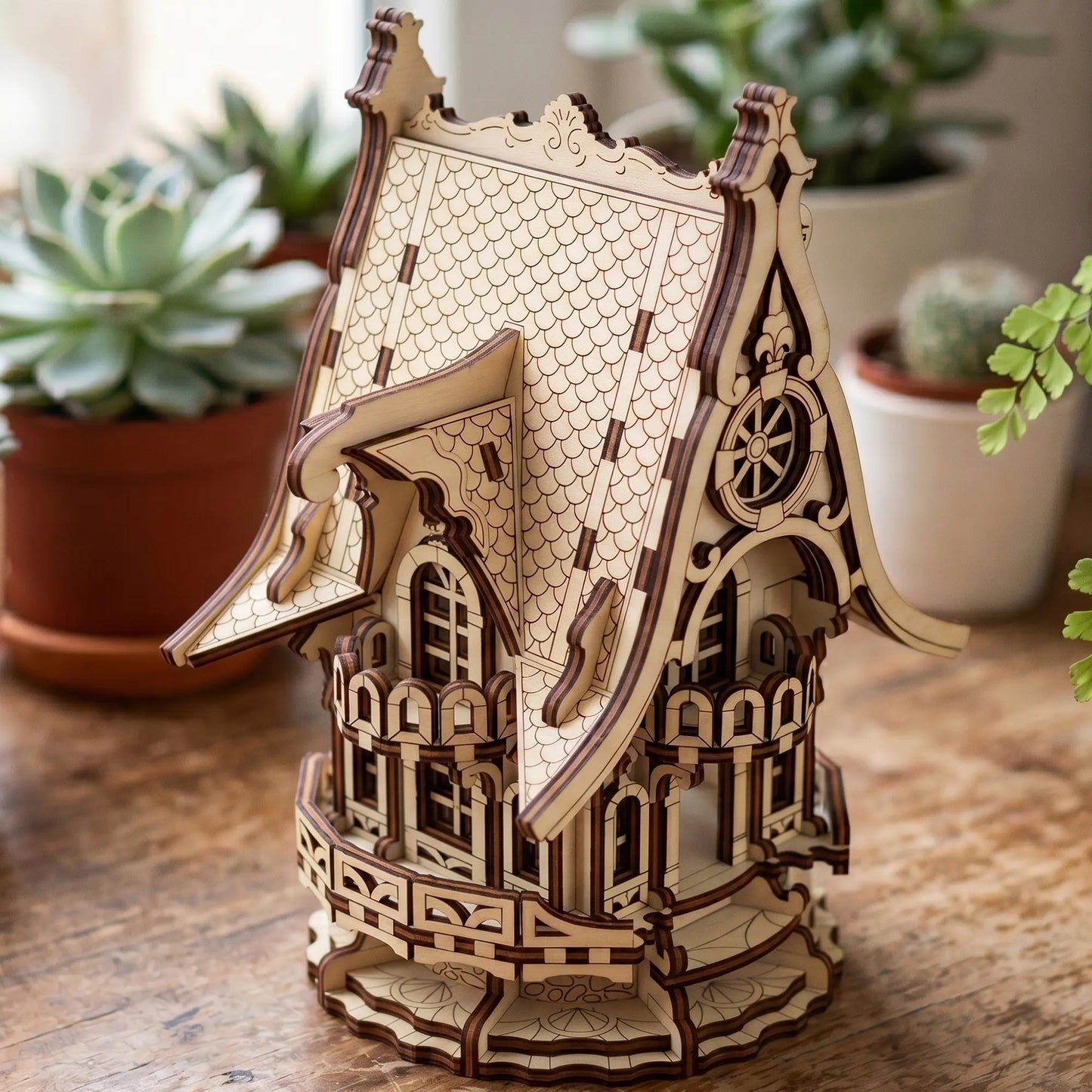 A 3D wooden model of a Victorian-style house with intricate laser-cut details, displayed on a wooden tabletop with potted plants in the background.