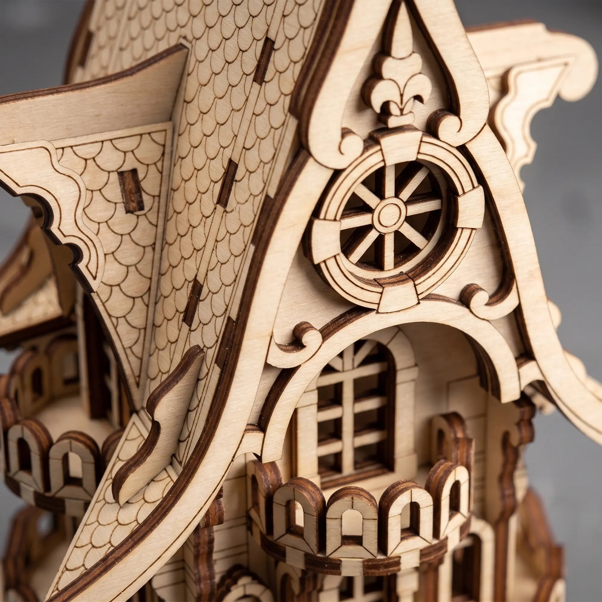 Close-up of a laser-cut wooden model of a fairy-tale house, showing detailed carved wood textures, a scalloped roof, and ornate architectural features against a soft gray background.