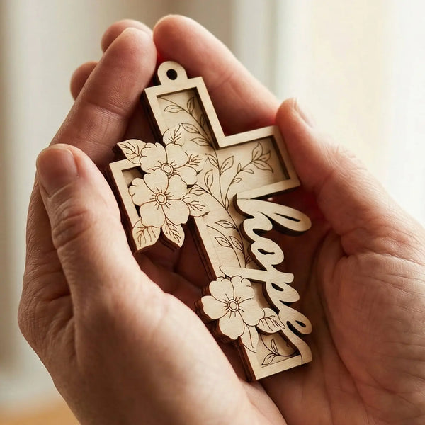 Hand holding a wooden laser cut cross ornament with floral designs and 'hope' engraved.