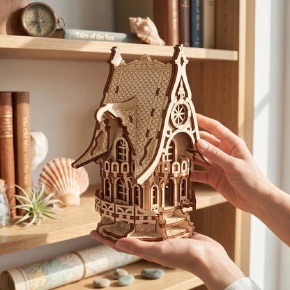 A person's hands holding a small wooden house model with intricate architectural details, set against a warm, sunlit background of wooden shelves filled with books and seashells.