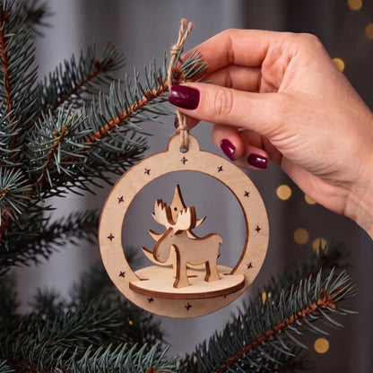 Hand hangs 3D laser cut plywood deer and tree ornament inside a star-cutout hoop on pine branches with lights.