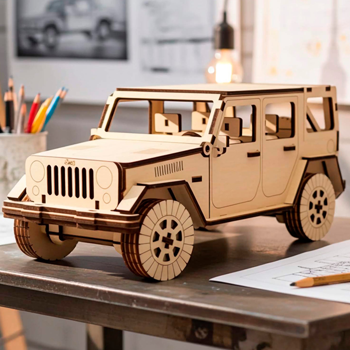 Front three-quarter view of a laser cut wooden plywood SUV model on a desk with pencils and sketches. Showcases intricate interior seating and exterior door handles. The natural wood grain is accented by dark brown laser-engraved lines.