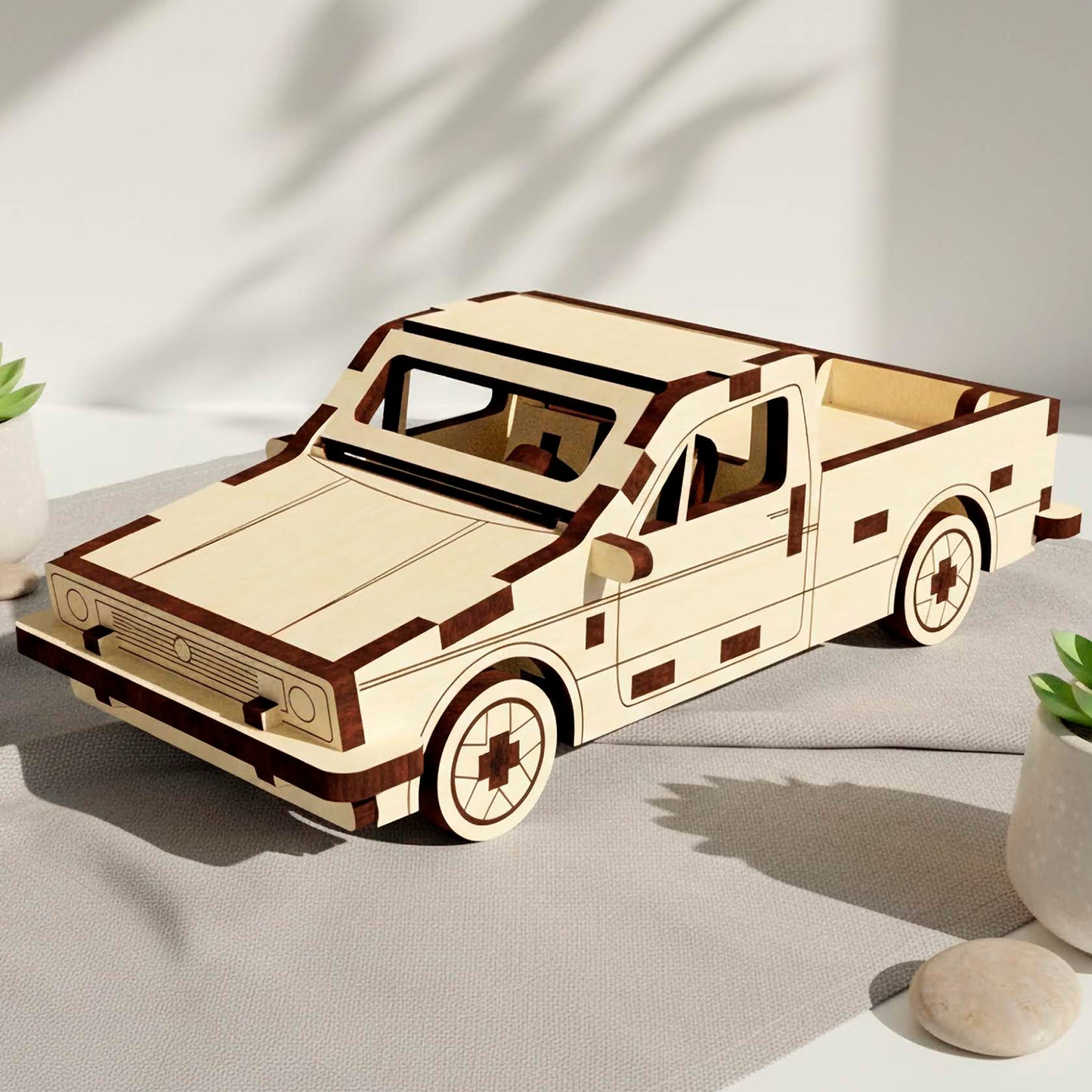 Laser cut wooden plywood pickup truck model with dark brown trim accents. The 3D puzzle car is displayed on a grey fabric surface with small succulents and smooth river stones under soft, leafy shadows on a white wall.