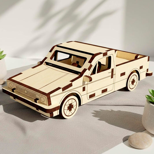 Laser cut wooden plywood pickup truck model with dark brown trim accents. The 3D puzzle car is displayed on a grey fabric surface with small succulents and smooth river stones under soft, leafy shadows on a white wall.