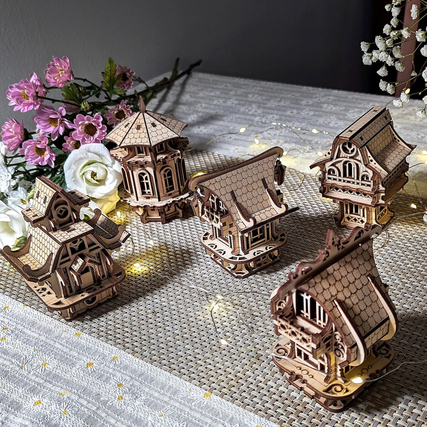 A collection of five miniature laser cut plywood buildings displayed next to a beautiful pink flower bouquet indoors.