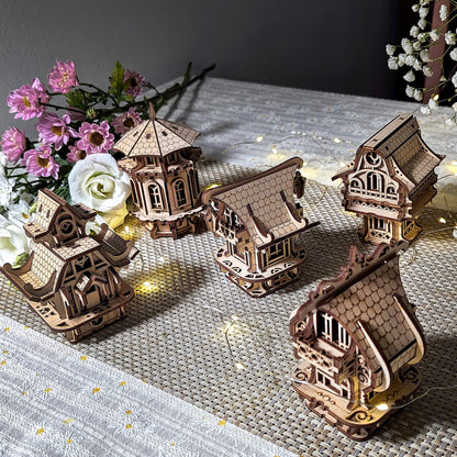 A collection of five miniature laser cut plywood buildings displayed next to a beautiful pink flower bouquet indoors.