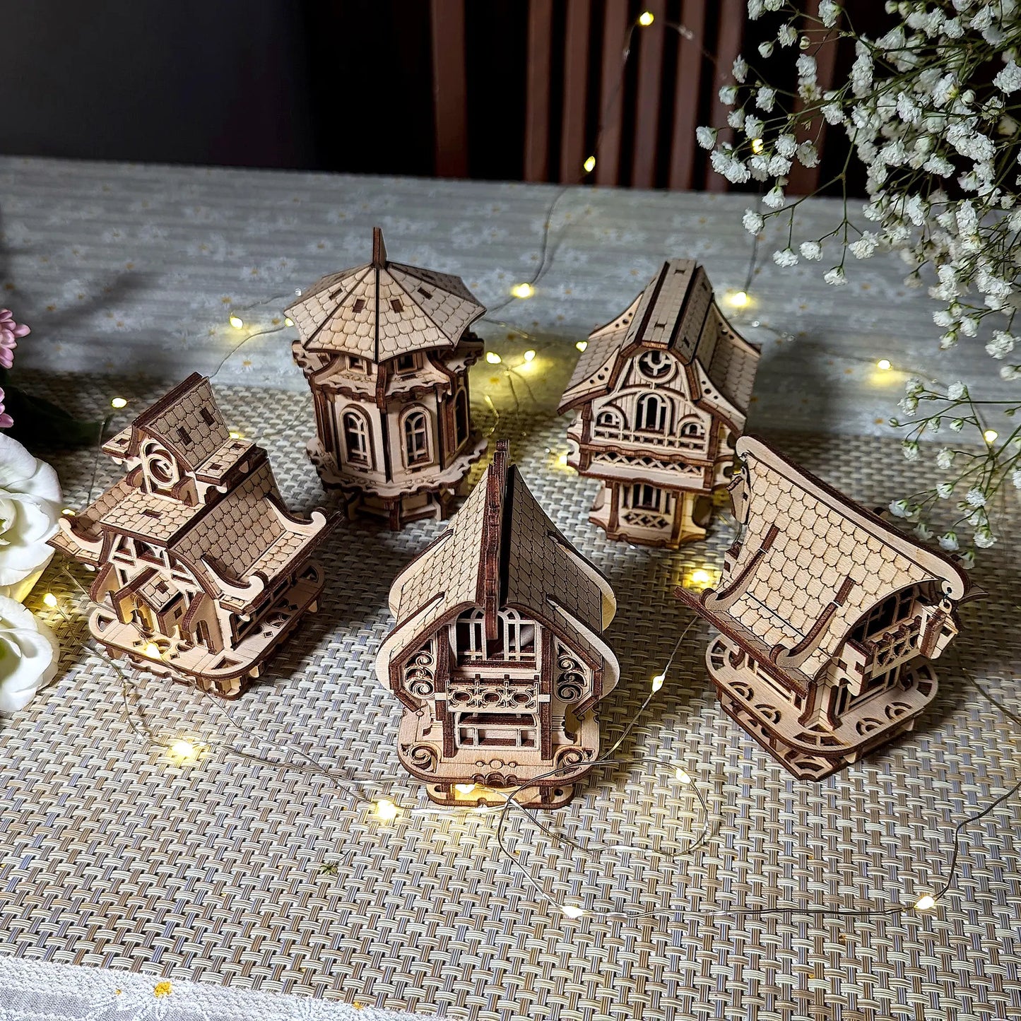 Top view of five diverse laser cut plywood village houses arranged together on a tablecloth with festive light string.