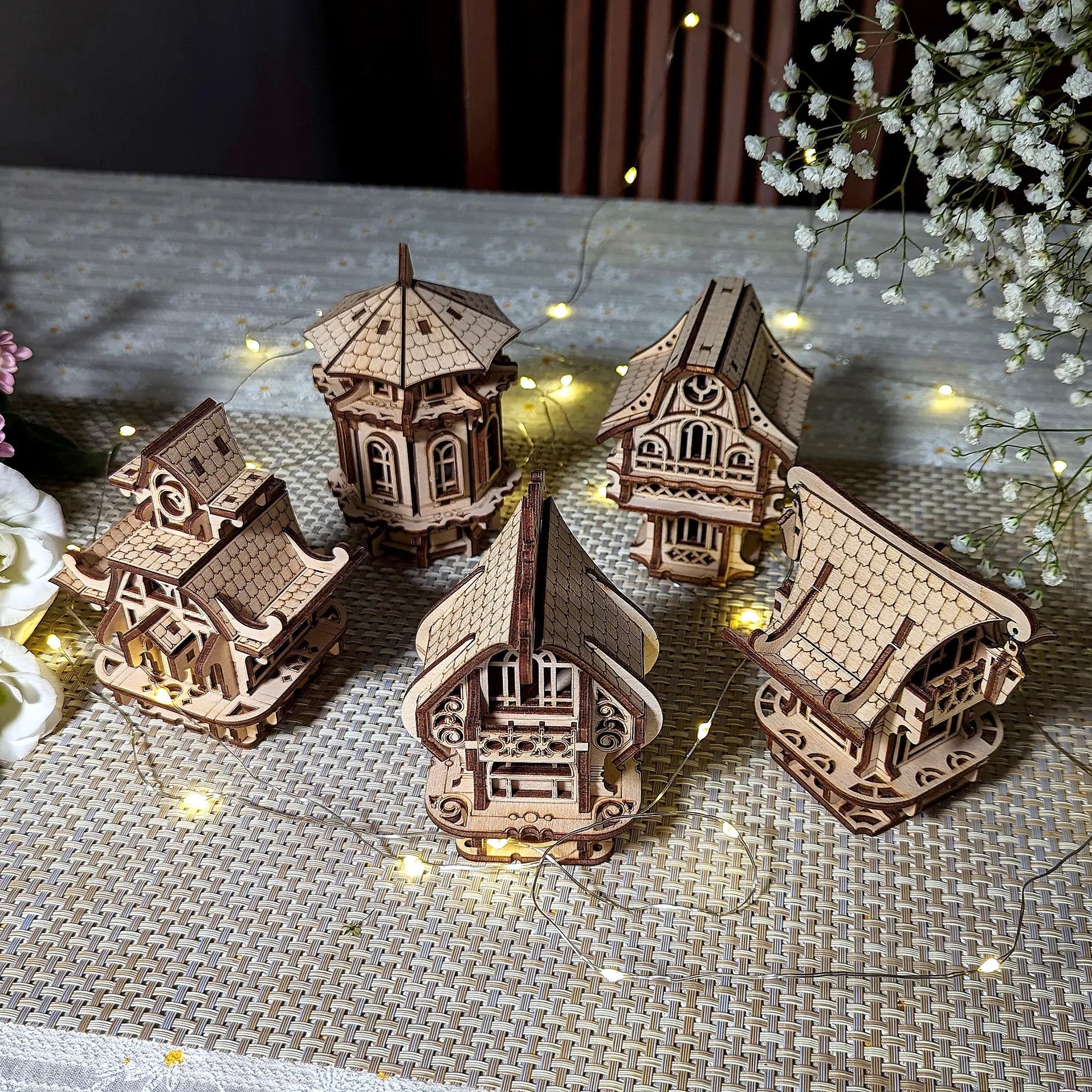 Top view of five diverse laser cut plywood village houses arranged together on a tablecloth with festive light string.