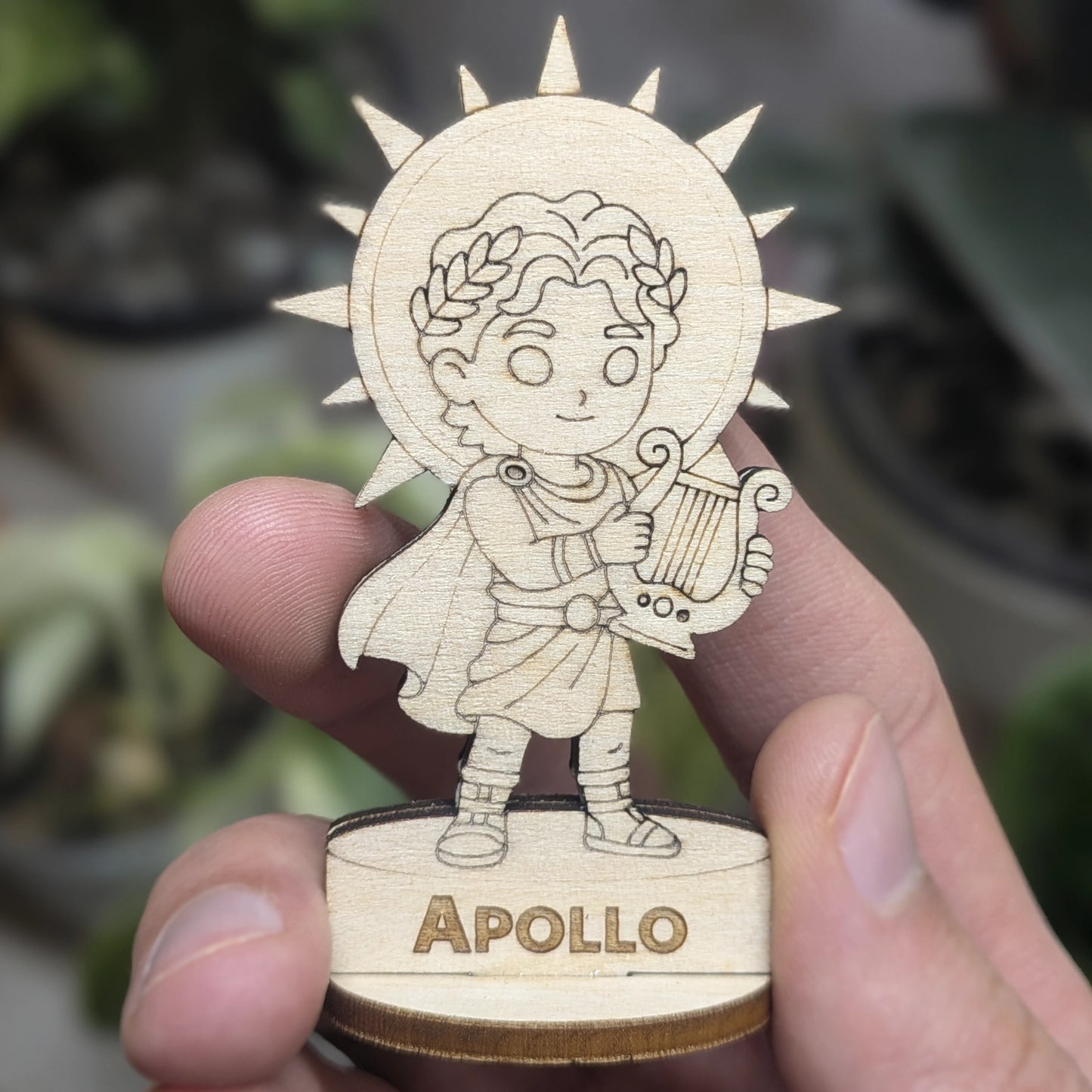 Hand holds a laser cut plywood Apollo for scale. Detailed front view shows the engraved lyre, laurel wreath, and wood robes