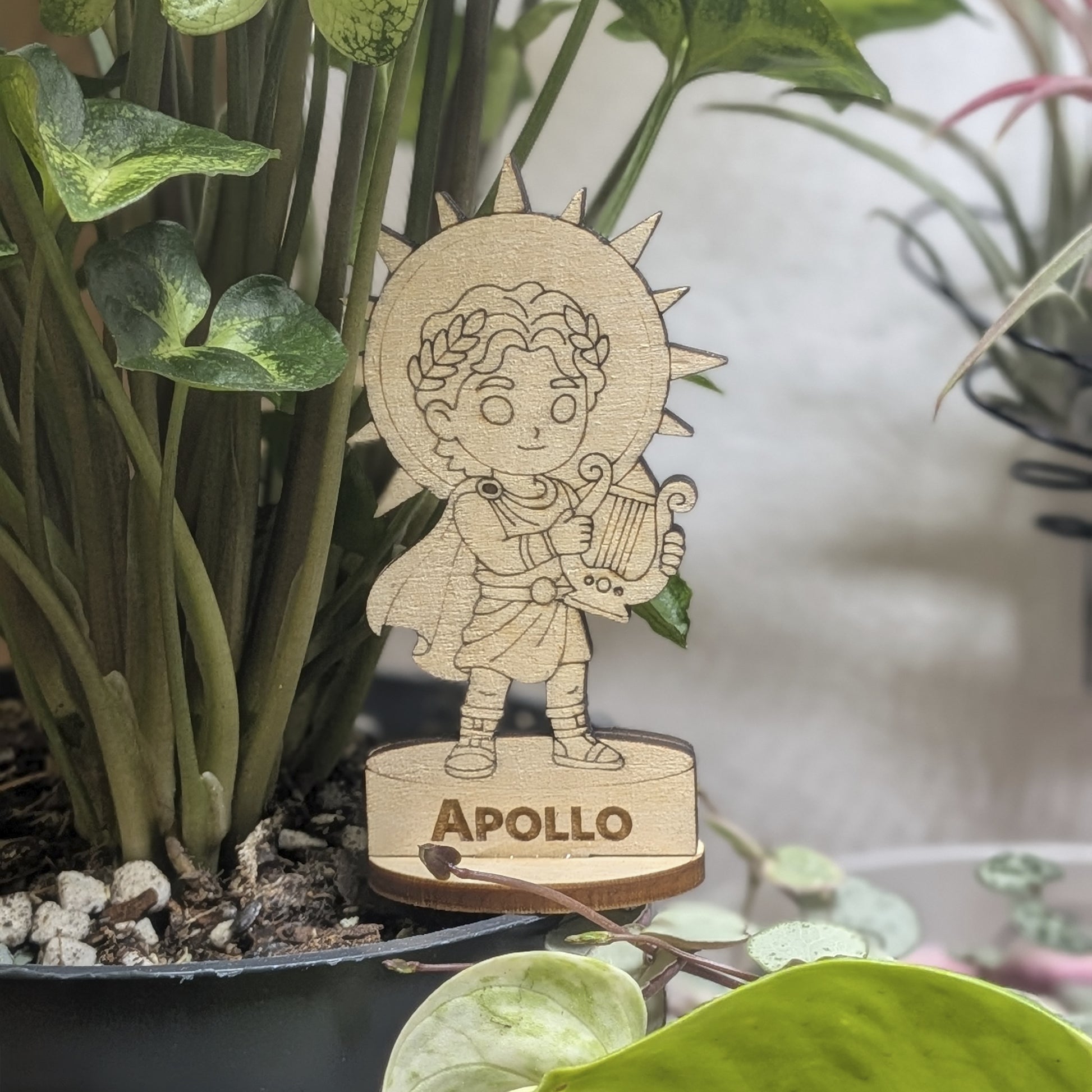 Laser cut plywood Apollo figurine sits in a plant pot. Features an engraved lyre, laurel crown, and robes on a wood base