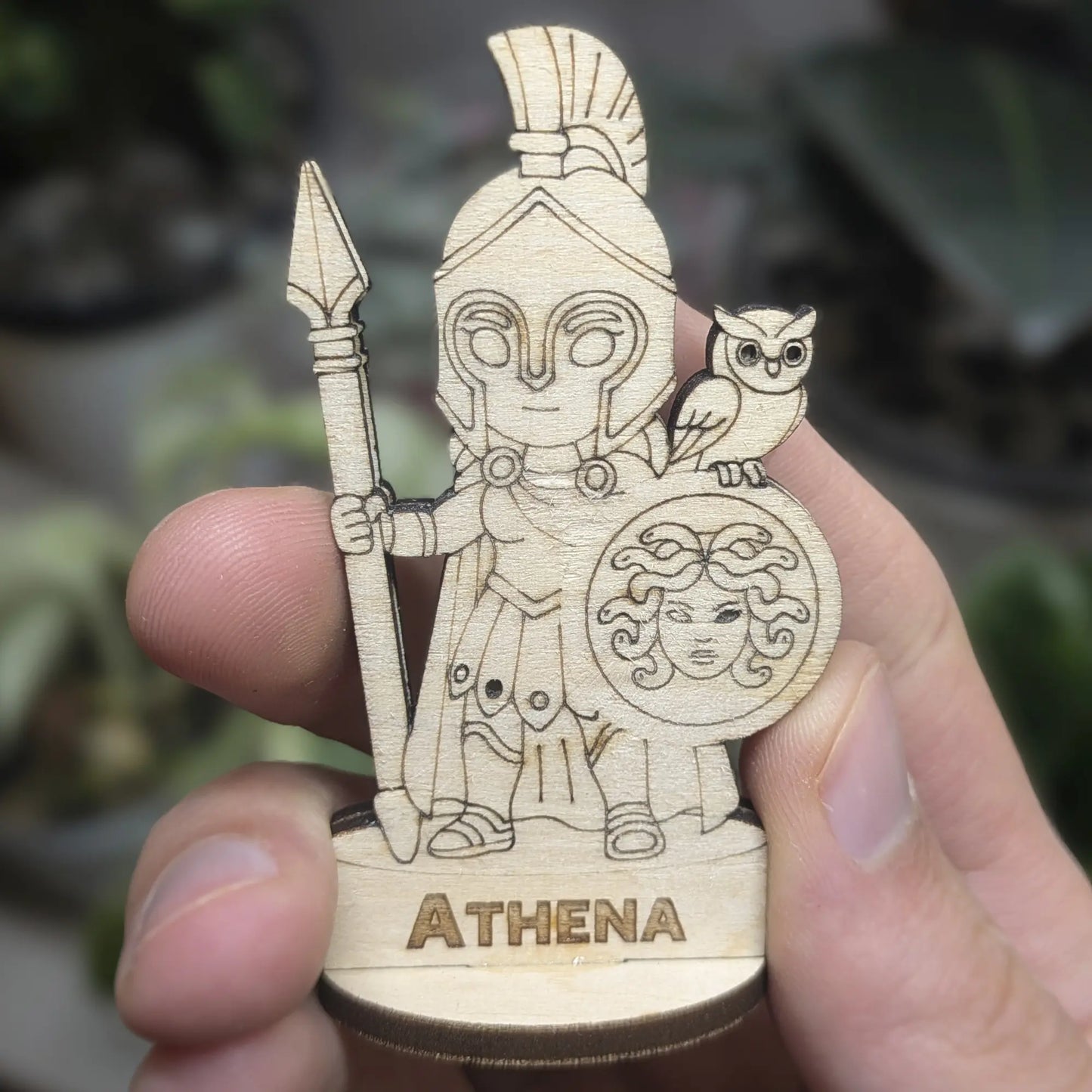 Hand holds an Athena goddess figurine of laser cut plywood. Shows the detailed helmet, spear, and Aegis owl shield at a tiny scale